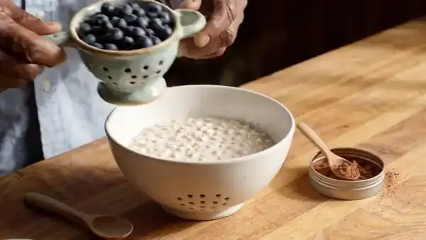 Hands tipping fresh blueberries into a bowl of oatmeal and stirring in a spoonful of cocoa