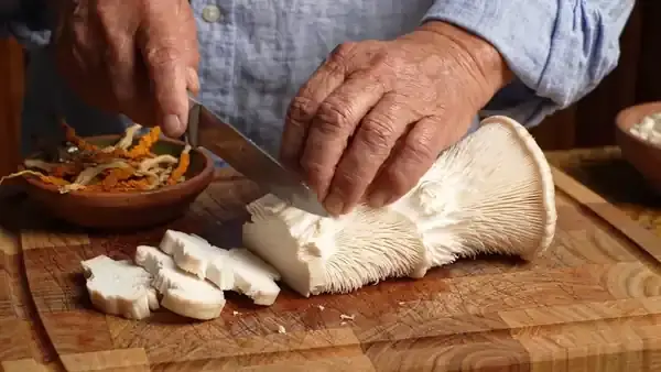 Hands slicing a fresh lion's mane mushroom into thick pieces on a walnut chopping board